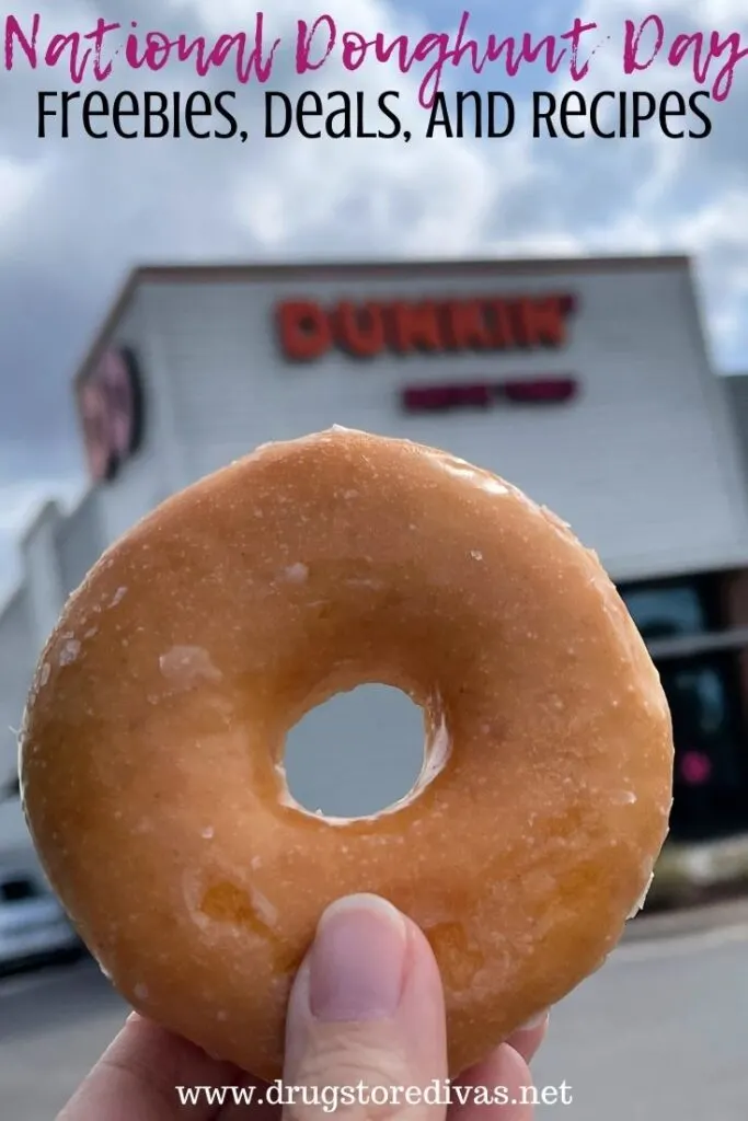 A hand holding a doughnut in front of a Dunkin' with the words "National Doughnut Day Freebies, Deals, And Recipes" digitally written above it.