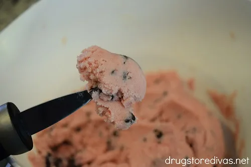 A melon baller scoop with pink dough on it over a bowl of pink dough.