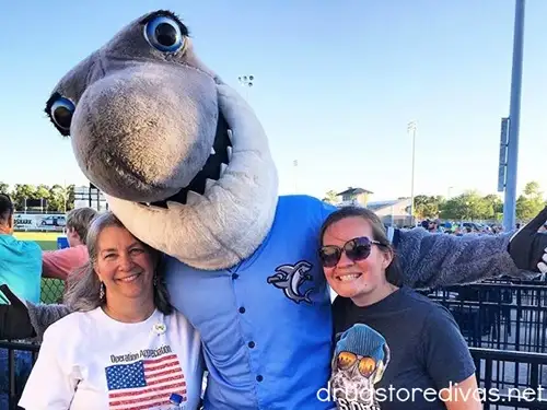 Two women standing at a baseball stadium with a shark mascot between them.