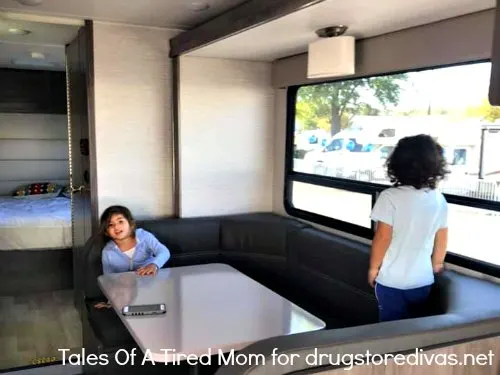 Two kids at the table in an RV, one is sitting and one is standing and looking out the window.
