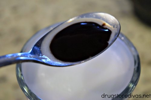 A spoon with chocolate syrup above a glass with a white liquid in it.