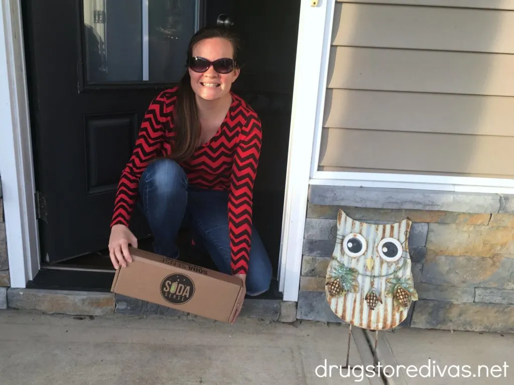 Smiling girl in a red and black chevron shirt, kneeling out an open front door, picking up a box with the words "Soda Sense". An owl lawn ornament is also in the photo.