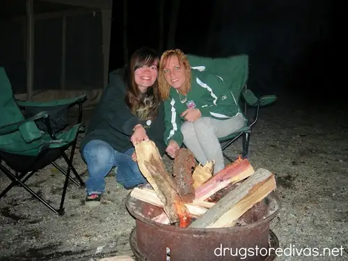 Two women sitting behind a firepit with a lot of firewood in it.