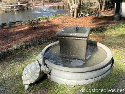 A water sculpture with a stone turtle on it in front of a pond with a water fountain in it.