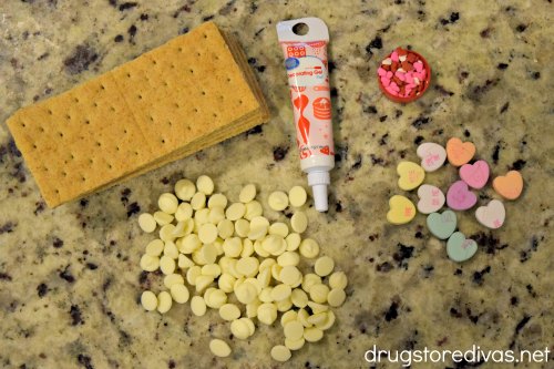 Graham crackers, white chocolate chips, red icing gel, heart-shaped sprinkles, and Conversation Hearts on a counter.