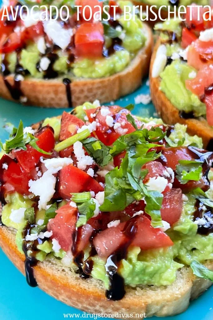 Three pieces of mini avocado toast, with mashed avocado, chopped tomatoes, balsamic glaze, feta cheese, and chopped cilantro on top on a blue tray with the words "Avocado Toast Bruschetta" digitally written on top.