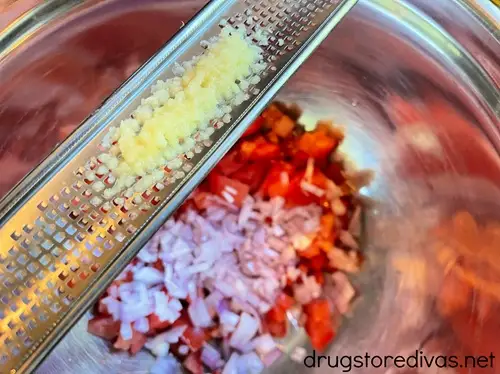 Garlic grated on a microplane grater above a silver bowl with diced tomato and shallot in it.