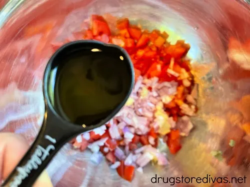 A black measuring spoon with a liquid in it above a silver bowl with diced tomato and shallot in it.