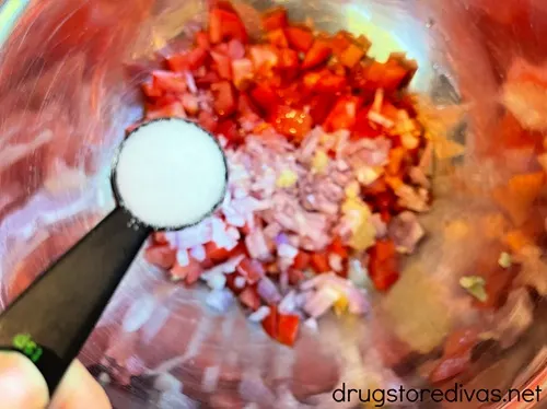 A black measuring spoon with salt in it above a silver bowl with diced tomato and shallot in it.