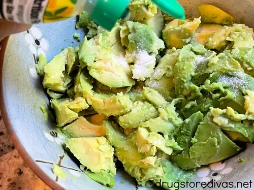 A squeeze bottle of lemon juice in a measuring spoon above the inside of avocado in a blue bowl.