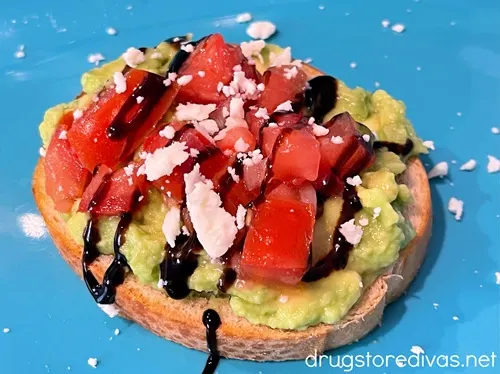Crumbled feta cheese, drizzles of balsamic glaze, diced tomatoes, and avocado mash on a small toast on a blue tray.