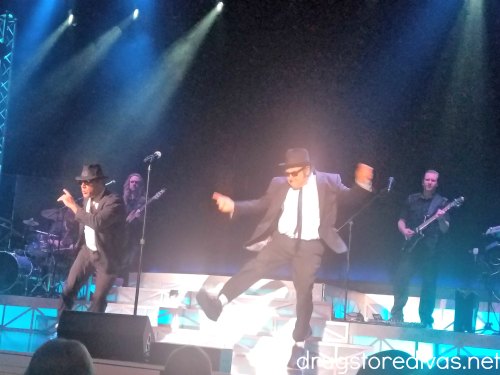 Two men, dressed as the Blues Brothers, dancing on stage. Two men in the back play guitar and drums.