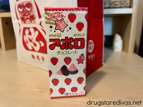A box of Meiji Apollo Strawberry Chocolates on a shelf with a red and white bag with a daruma on it behind it.
