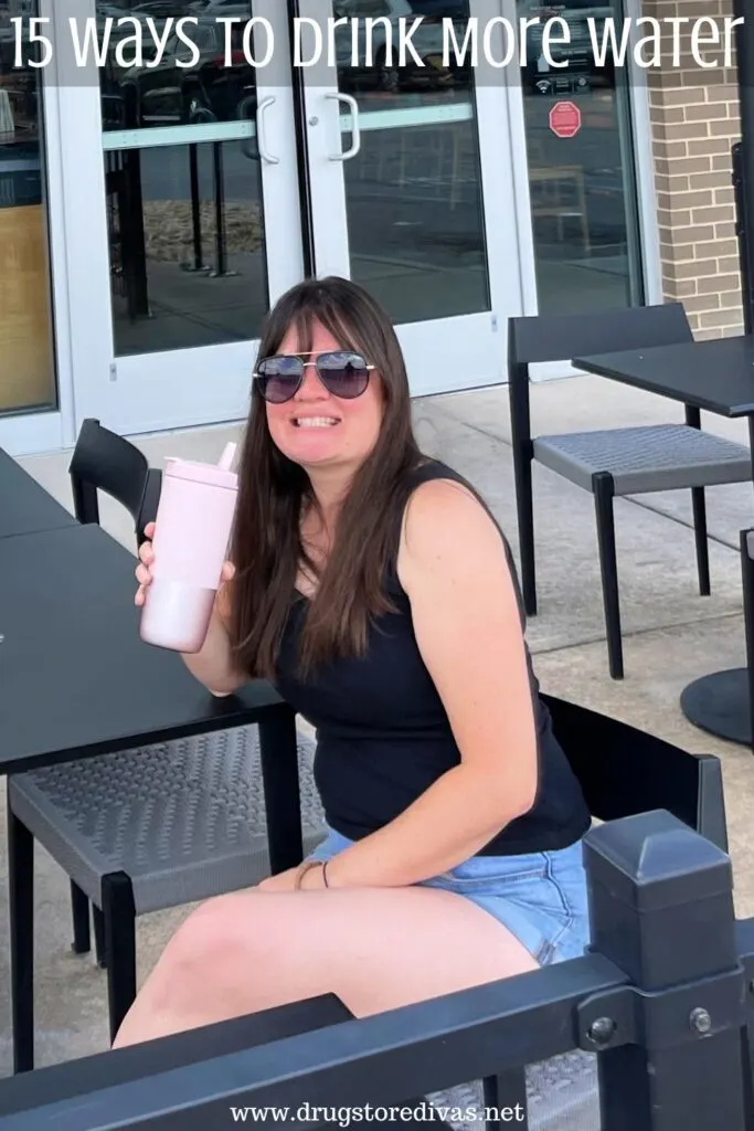 A woman in a black top and jean shorts and sunglasses, sitting at a table, holding a pink water bottle, with the words "15 Ways To Drink More Water" digitally written above her.