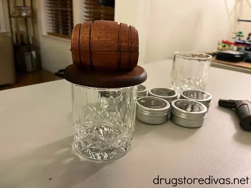 A whiskey smoker kit, which includes two glasses, four metal containers, an a whiskey barrel on top of one of the cups, on a table.