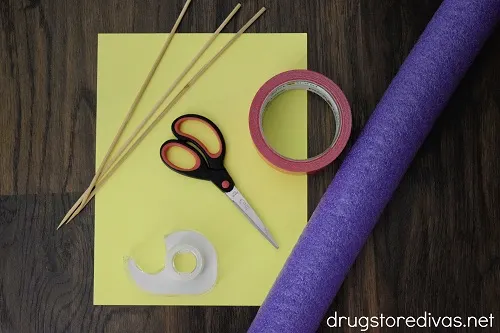 Skewers, tape, scissors, duct tape, a pool noodle, and a yellow piece of paper on a wooden background.