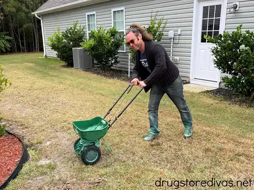 A man in a black sweater and jeans pushing a green seed spreader.
