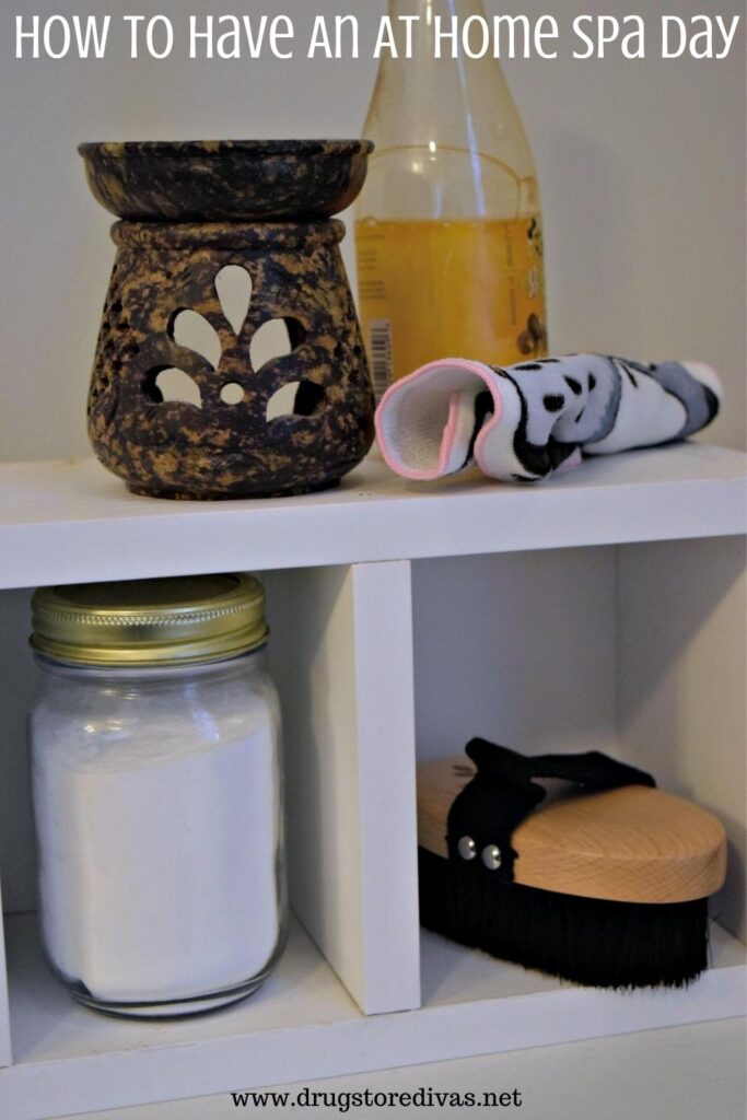 A candle, bubble bath, a towel, body powder, and a body brush on a shelf with the words "How To Have An At Home Spa Day" digitally written on top.