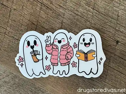 A sticker of three ghosts, one is holding coffee, one is in a pink jacket, and one is holding a book.