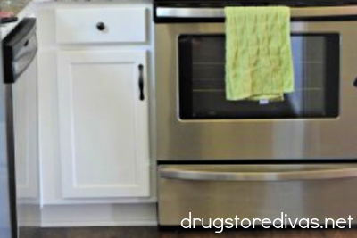 A kitchen oven with a green towel hanging on the door with a white cabinet next to it.