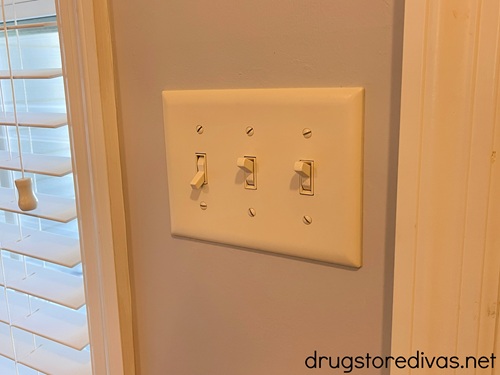 A triple light switch on a wall with a window off to the side.