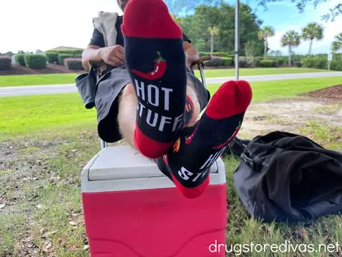 A man sitting in a chair with his feet on a red and white cooler while wearing a pair of socks that say hot stuff at the bottom.