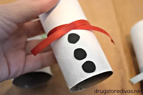 A white cardboard tube with a red ribbon in the middle and three black circles down the bottom half.