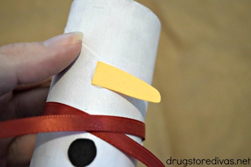 A white cardboard tube with an orange carrot-shaped nose, a red ribbon, and one black circle.