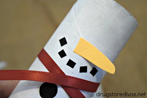 A white cardboard tube with a carrot-shaped nose, five black squares to make a smile, a maroon ribbon, and a black circle. 