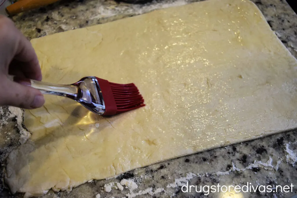 A pastry brush spreading butter on cinnamon roll dough in a rectangle on a countertop.