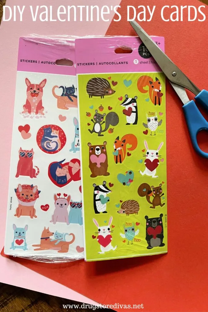 Two packs of Valentine's Day stickers a a pair of blue handled scissors on top of a pink and a red piece of construction paper with the words "DIY Valentine's Day Cards" digitally written on top.