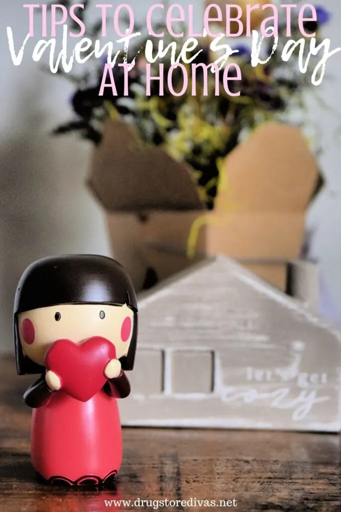 A wooden doll holding a heart, a wooden home that says let's get cozy, and flowers with the words "How To Celebrate Valentine's Day At Home" digitally written on top.