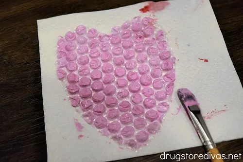 A bubble wrap heart painted pink on top of a napkin, next to a paint brush.