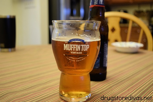 A muffin top beer glass with a beer bottle behind it on a table.