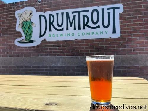 A beer sitting on a wooden table with a large DrumTrout Brewing Company sign behind it, in Wilmington, NC.