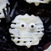 Oreo cookies decorated with white almond bark and candy eyes to look like a mummy, on black paper shred, with the words 