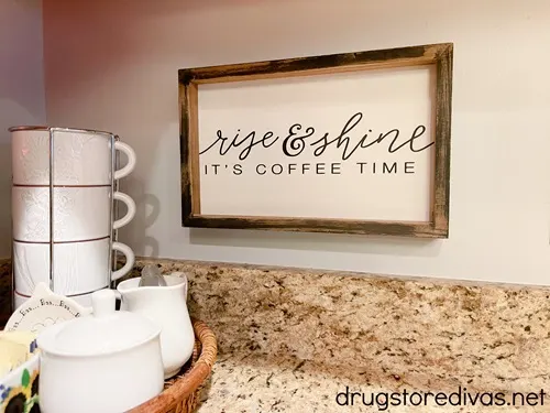 A wooden sign that says rise & shine it's coffee time next to a set of coffee mugs and sugar dish.