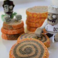 Homemade black and orange swirl pinwheel cookies, with two Halloween zombie toys, on a tray and the words 