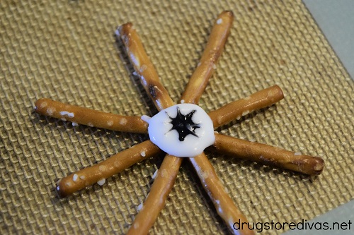 Black dot of decorating gel, made to look like a spider, in the middle of a white almond bark circle that's in the middle of eight pretzels.