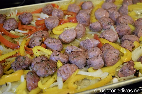 Sliced onions, peppers, garlic, and beef sausage rounds, topped with seasoning, on a sheet pan.