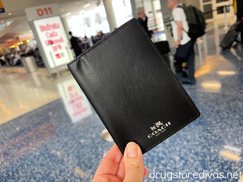 Fingers holding a black Coach-branded passport holder in an airport.