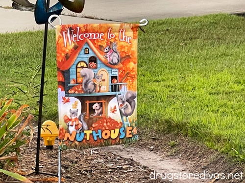 A garden flag with a house and squirrels on it that says Welcome To The Nut House.