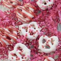 White chocolate-covered pretzels with sprinkles and red chocolate on them, on top of pink paper shred, with the words 
