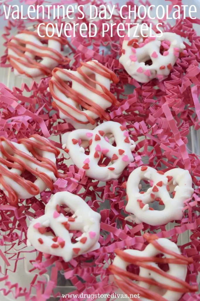 White chocolate-covered pretzels with sprinkles and red chocolate on them, on top of pink paper shred, with the words "Valentine's Day Chocolate Covered Pretzels" digitally written on top.