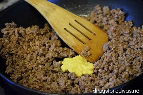 Ground beef and mustard in a pan with a bamboo spoon.