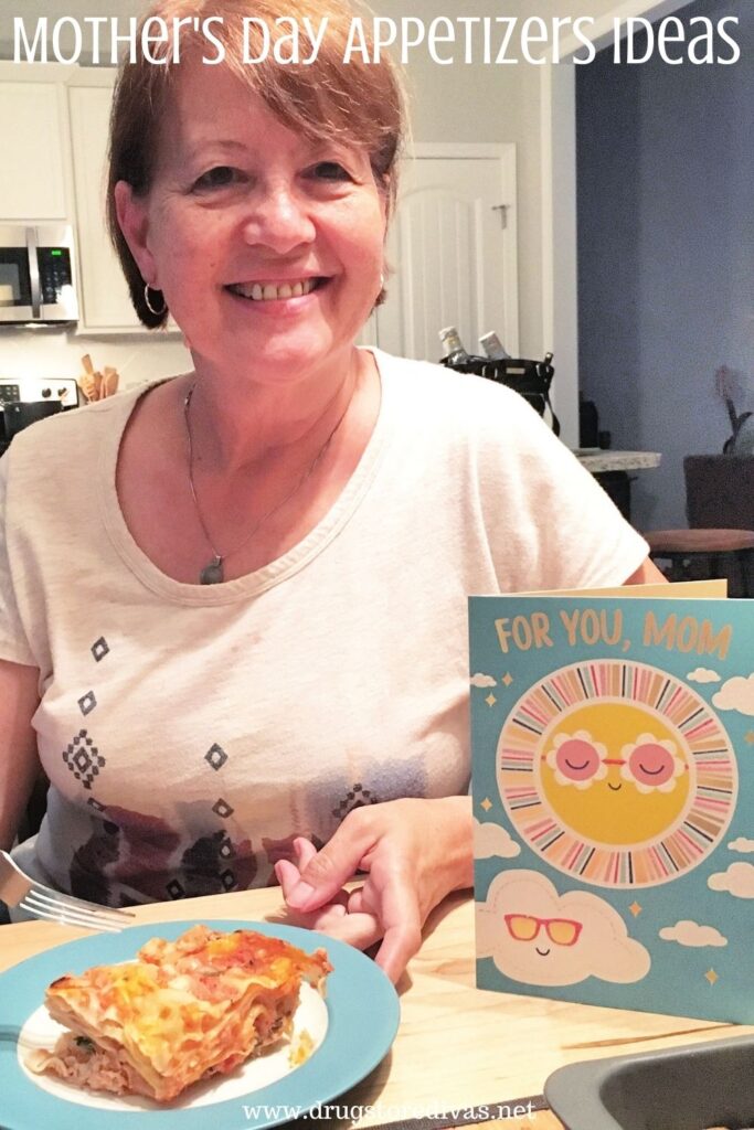 A woman sitting behind a plate of food in a kitchen, next to a Mother's Day card, with the words "Mother's Day Appetizers Ideas" digitally written on top of her.