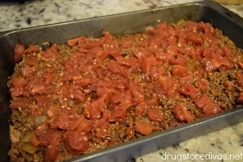 Diced tomatoes on top of ground beef spread out in a cake pan.