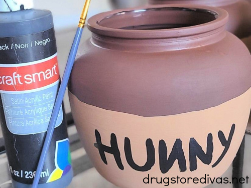 A two toned brown jar with the word "HUNNY" painted in black, next to a tube of black paint and a paint brush.