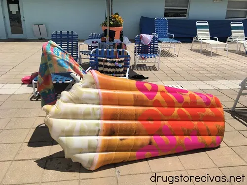 A Dunkin' iced coffee shaped pool float in front of pool chairs and a towel.