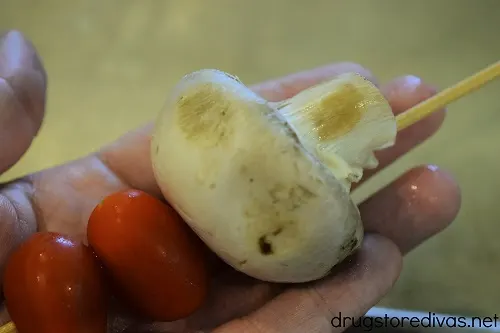 A hand holding a skewer with a mushroom and tomato on it.